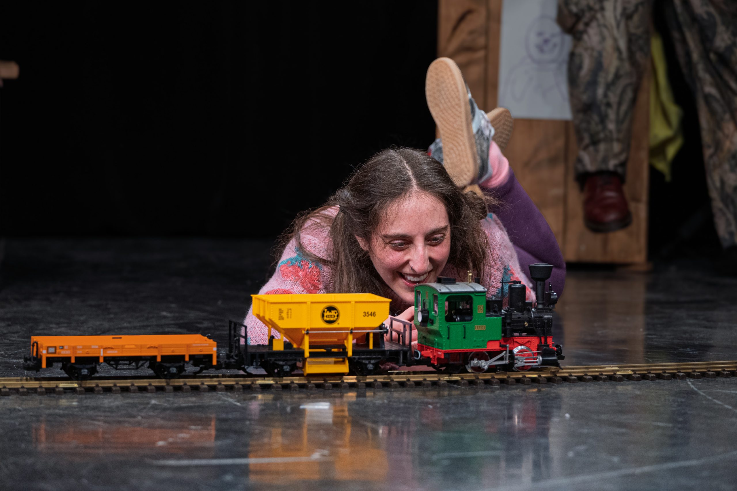 Kind liegt auf dem Boden und spielt mit einer Modellbahn mit grün-roter Lok und gelbem Wagen auf Schienen