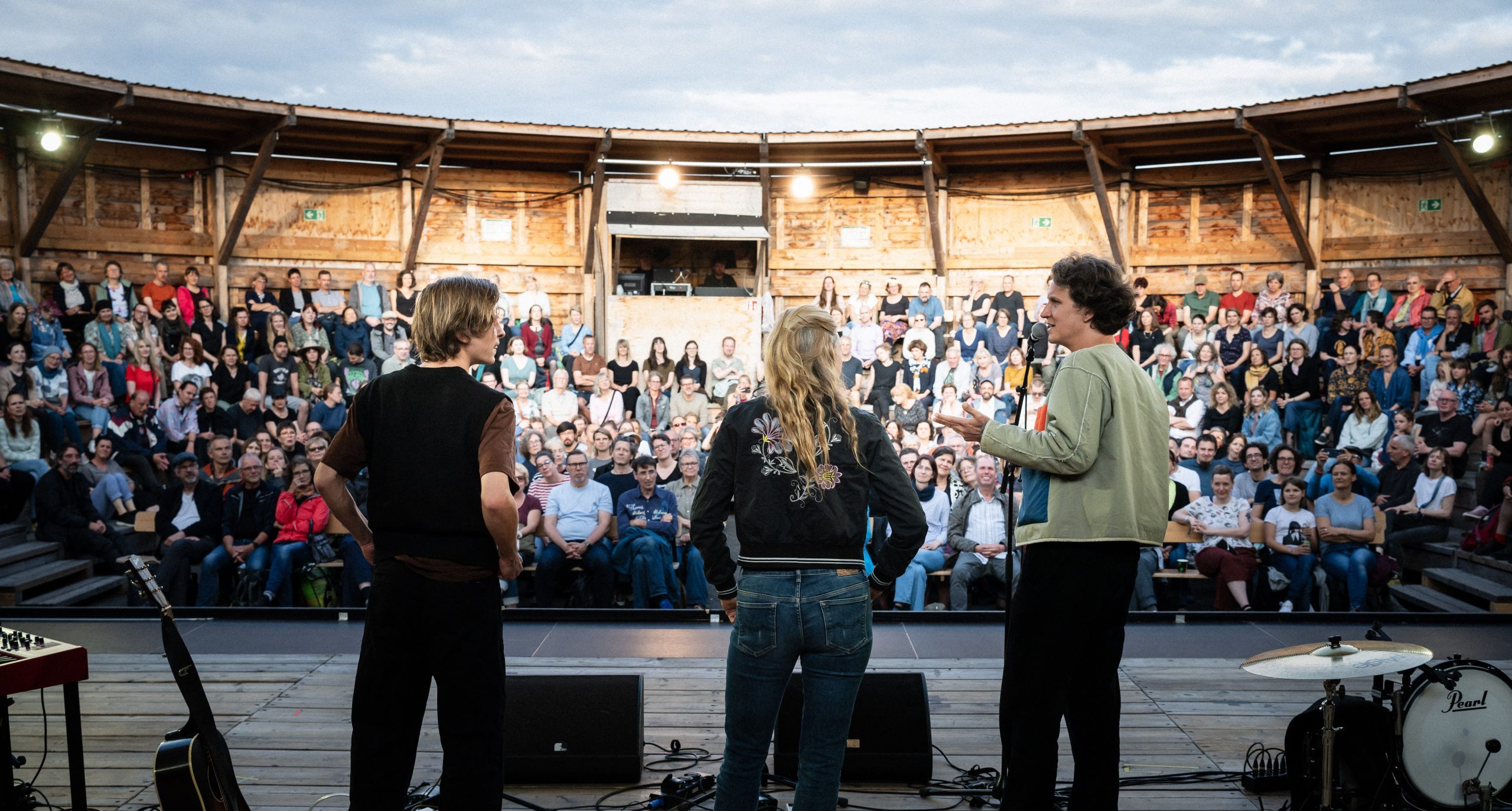 Drei Personen stehen auf einer Bühne vor einem Publikum in einem runden, hölzernen Theater