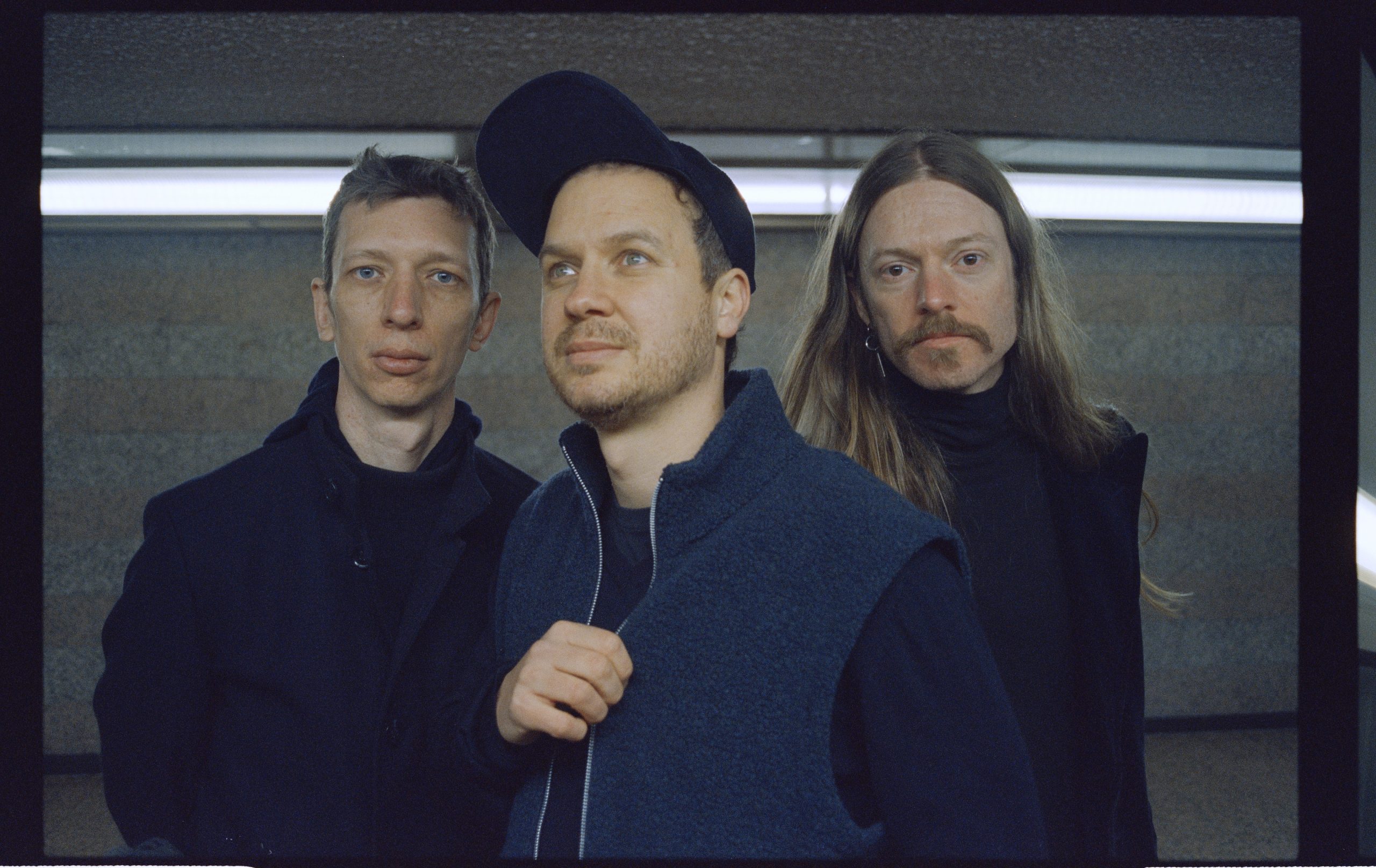 Drei Personen stehen nebeneinander vor einer Wand mit horizontalem Lichtstreifen, die mittlere Person trägt eine Kappe und eine Weste, die anderen zwei Personen tragen dunkle Kleidung