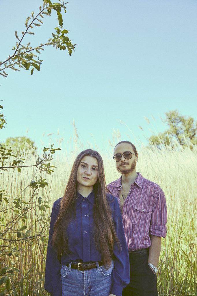 Zwei Personen stehen in einem Feld mit hohem Gras vor blauem Himmel, die vordere trägt ein dunkelblaues Hemd und Jeans, die hintere ein gestreiftes Hemd mit hochgekrempelten Ärmeln