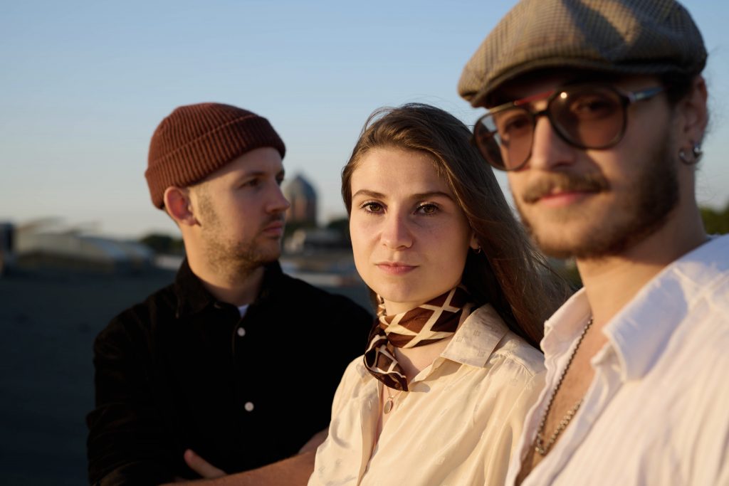 Drei Personen stehen im Freien bei Sonnenlicht, eine trägt eine braune Strickmütze, eine weitere ein helles Hemd mit einem gemusterten Halstuch, die dritte Person trägt ein weißes Hemd und eine Kappe.