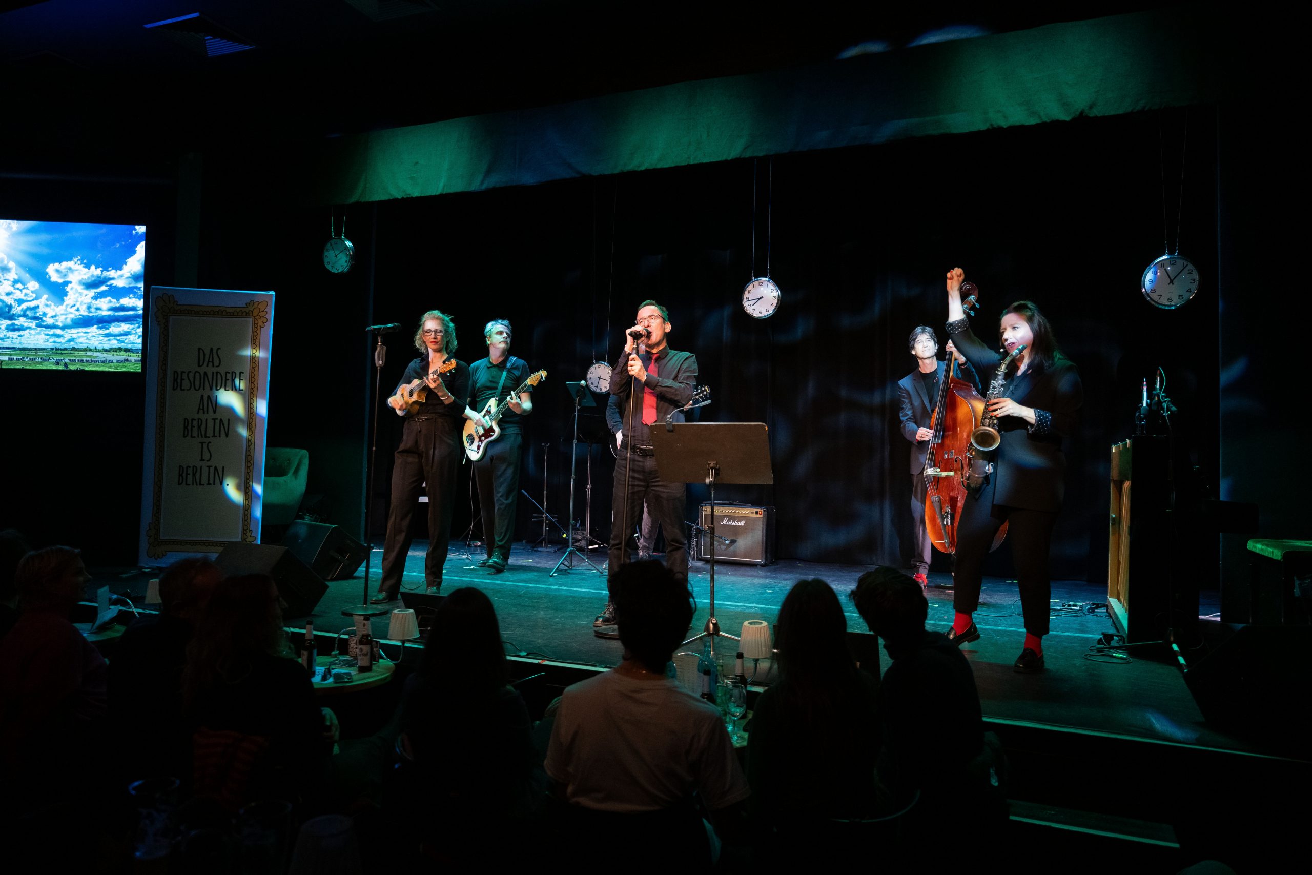 Band vor Publikum steht auf einer Bühne und spielt Musik.