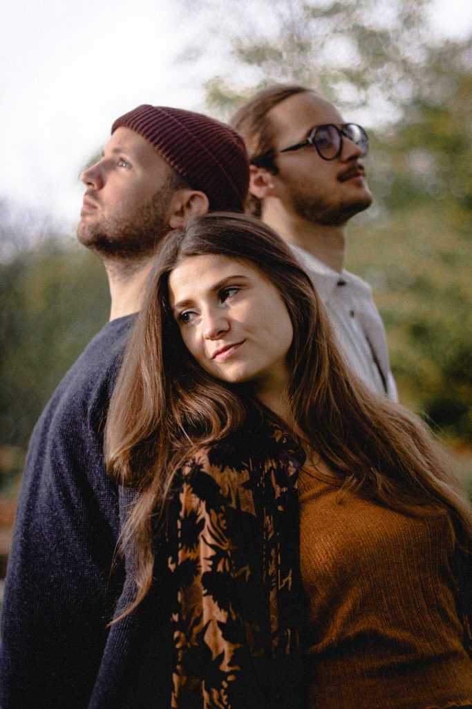 Drei Personen stehen im Freien vor unscharfem Hintergrund, eine trägt eine dunkelrote Mütze, eine weitere ein braunes Oberteil mit floralem Muster