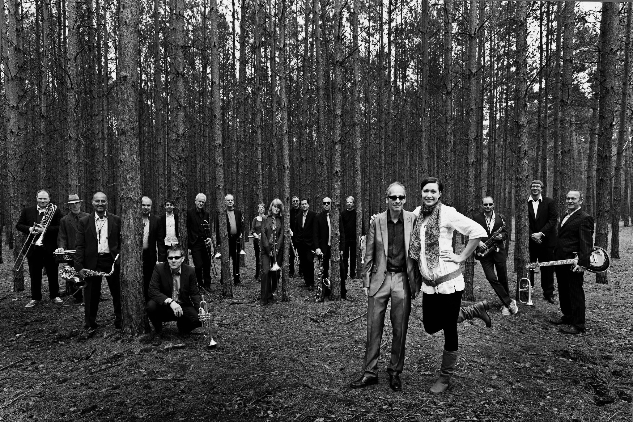 Eine große Gruppe an Menschen steht zwischen vielen Bäumen im Wald, während zwei Männer im Vordergrund stehen.