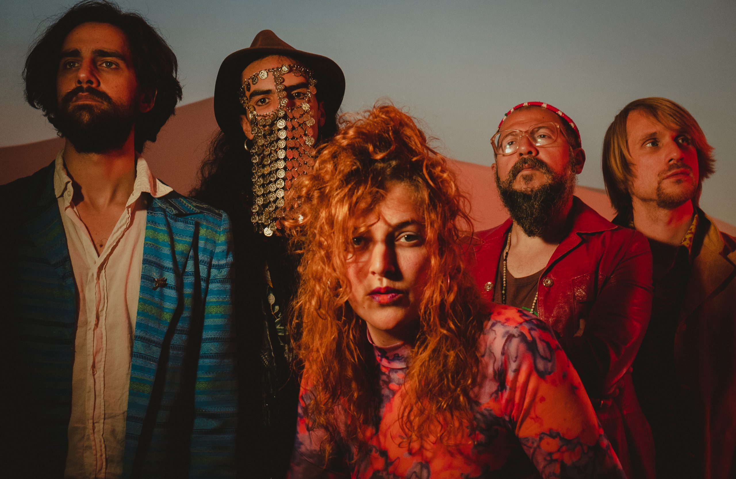 Eine Gruppe von 5 Personen in bunten Klamotten steht vor einem Wüstenhintergrund. Die einzige Frau steht im Vordergrund.