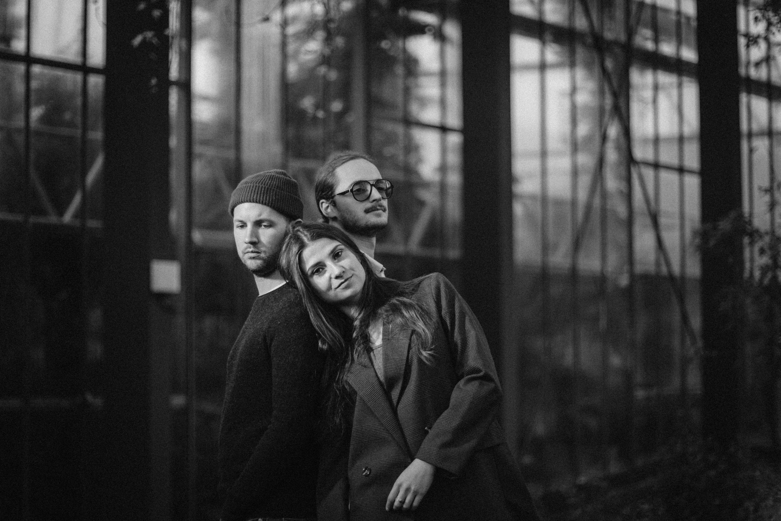 Drei Personen stehen eng beieinander vor einem großen Fenster mit Gitterrahmen in einem Innenraum