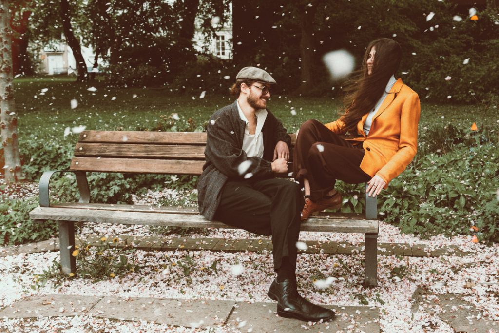 Zwei Personen sitzen auf einer Parkbank, eine trägt eine orangefarbene Jacke und schwarze Hose, die andere eine graue Mütze und dunkle Kleidung, umgeben von fallenden Blütenblättern.