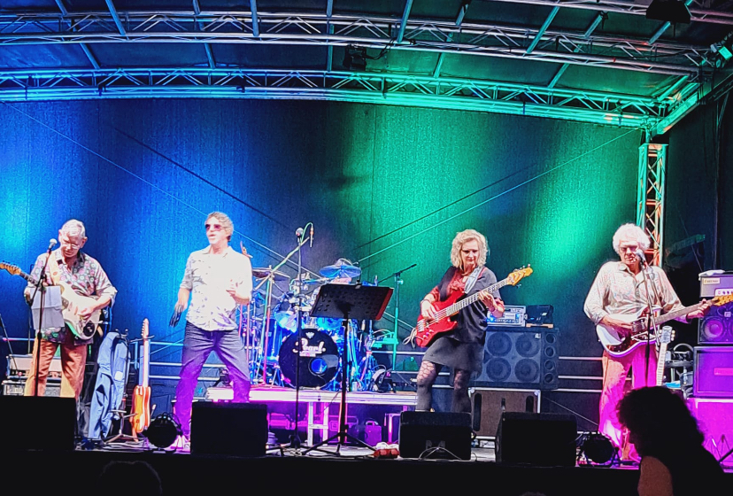 Vier Personen stehen auf einer bunt beleuchteten Bühne. Drei Personen spielen E-Gitarre oder Bass. Die vierte Person spielt ein Tamburin.