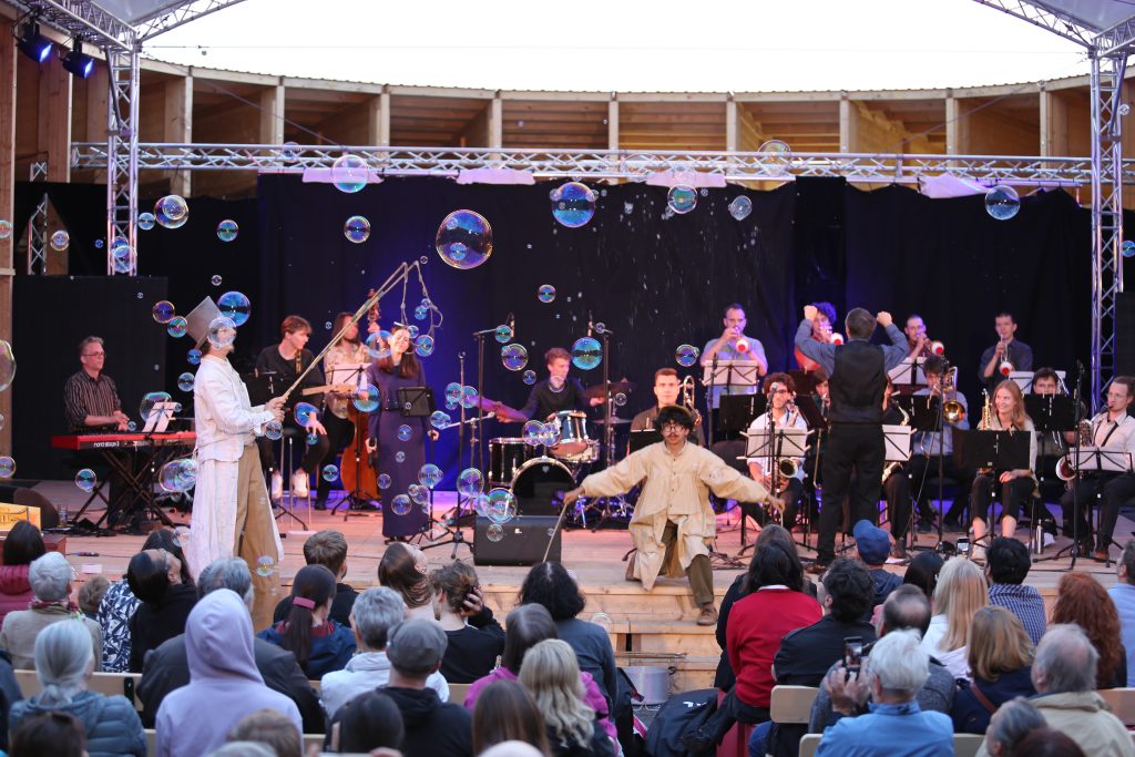 Big Band spielt auf einer hölzernen Open Air Bühne vor Publikum, während zwei Männer Tricks mit Seifenblasen auf der Bühne machen.