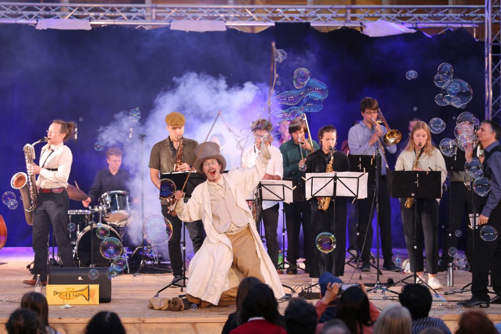 Big Band spielt auf einer Bühne vor Publikum, während ein Mann Tricks mit Seifenblasen auf der Bühne macht.