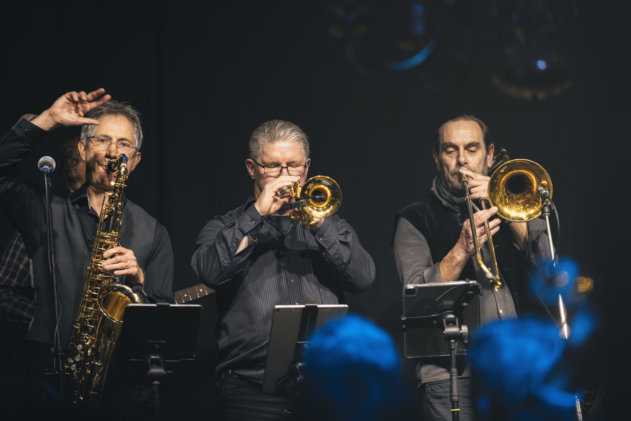 Drei Männer in schwarz spielen ihre jeweiligen Blasinstrumente.
