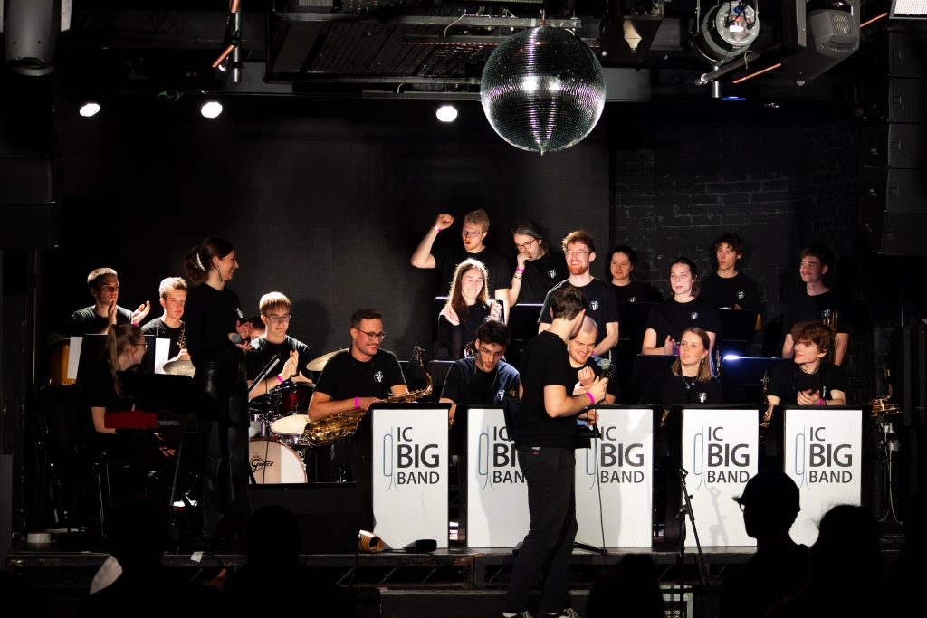 Big Band spielt in einem dunklen Raum von Publikum. An der Decke hängt eine Diskokugel.