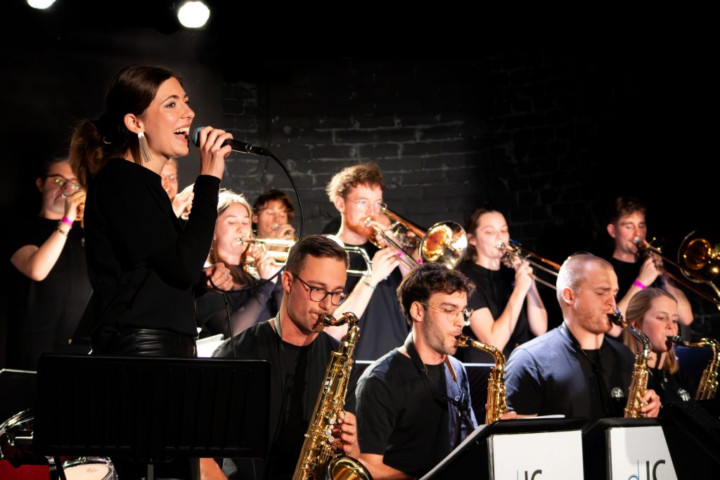 Big Band spielt in einem dunklen Raum von Publikum. Eine Sängerin steht an der linken Seite und singt.