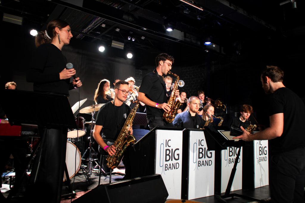 Big Band spielt in einem dunklen Raum von Publikum. Eine Sängerin steht an der linken Seite und singt, während ein Dirigent die Band dirigiert.