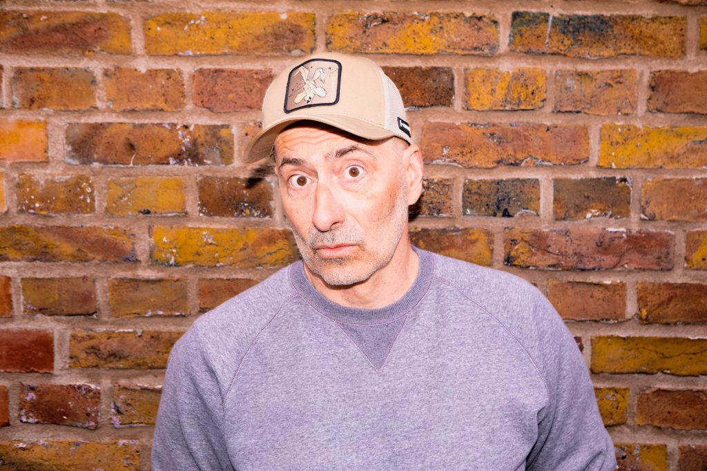 Ein Mann mit Kappe und grauen Pullover steht vor einer Backsteinwand.