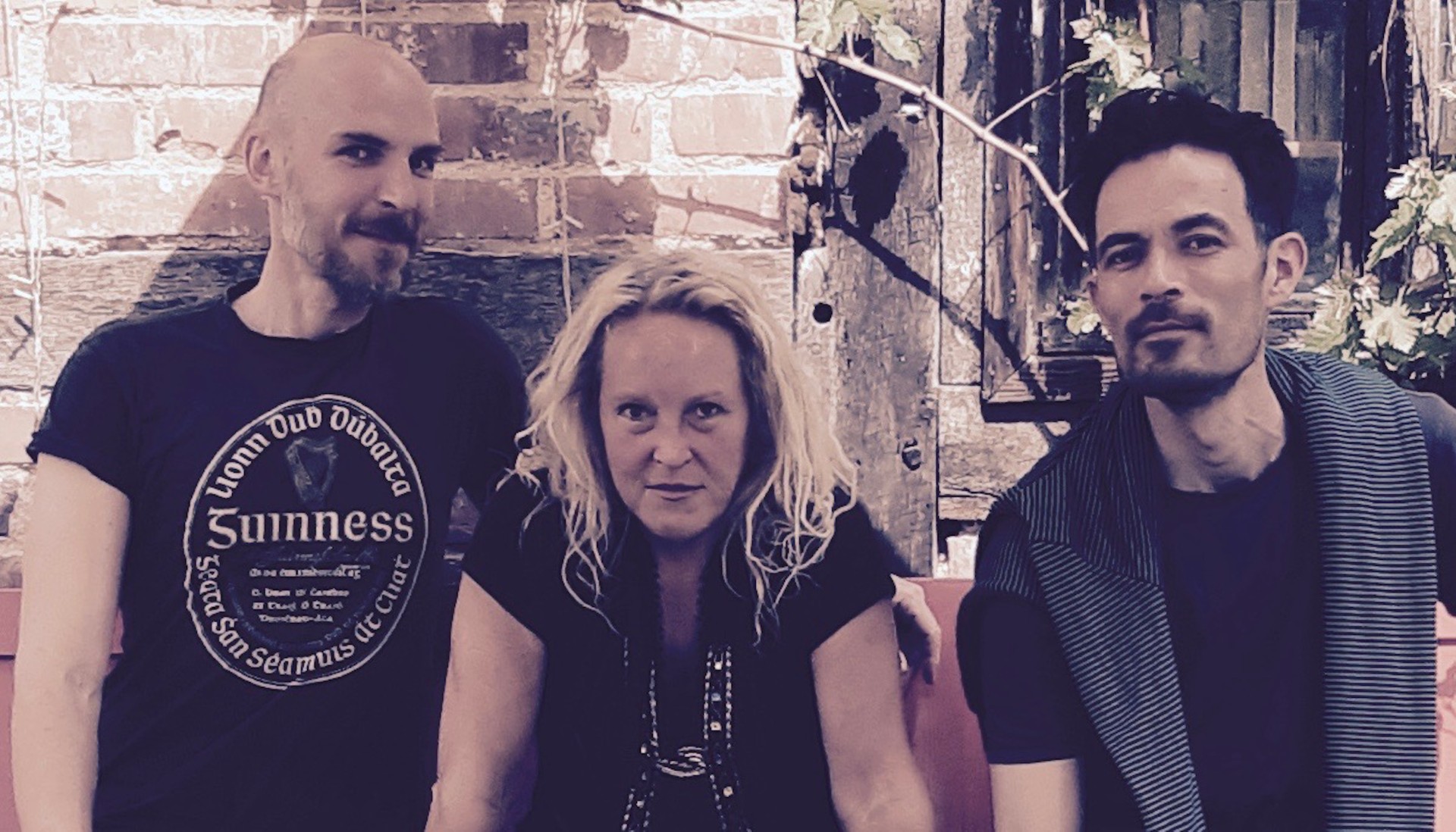 Drei Personen sitzen vor einer Backsteinmauer, die mittlere Person hat lange blonde Haare, die linke trägt ein schwarzes T-Shirt mit rundem Aufdruck, die rechte Person trägt ein gestreiftes Oberteil über einem schwarzen T-Shirt.