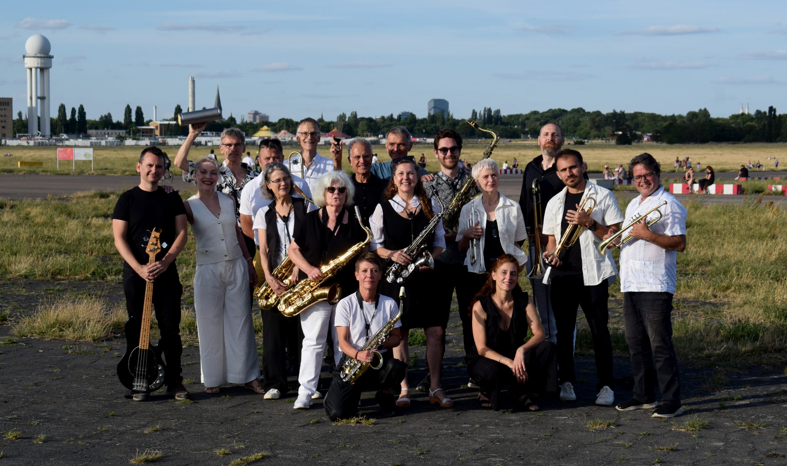 Gruppe von Menschen mit Blasinstrumenten und Bassgitarre steht auf offenem Gelände mit Gebäuden im Hintergrund