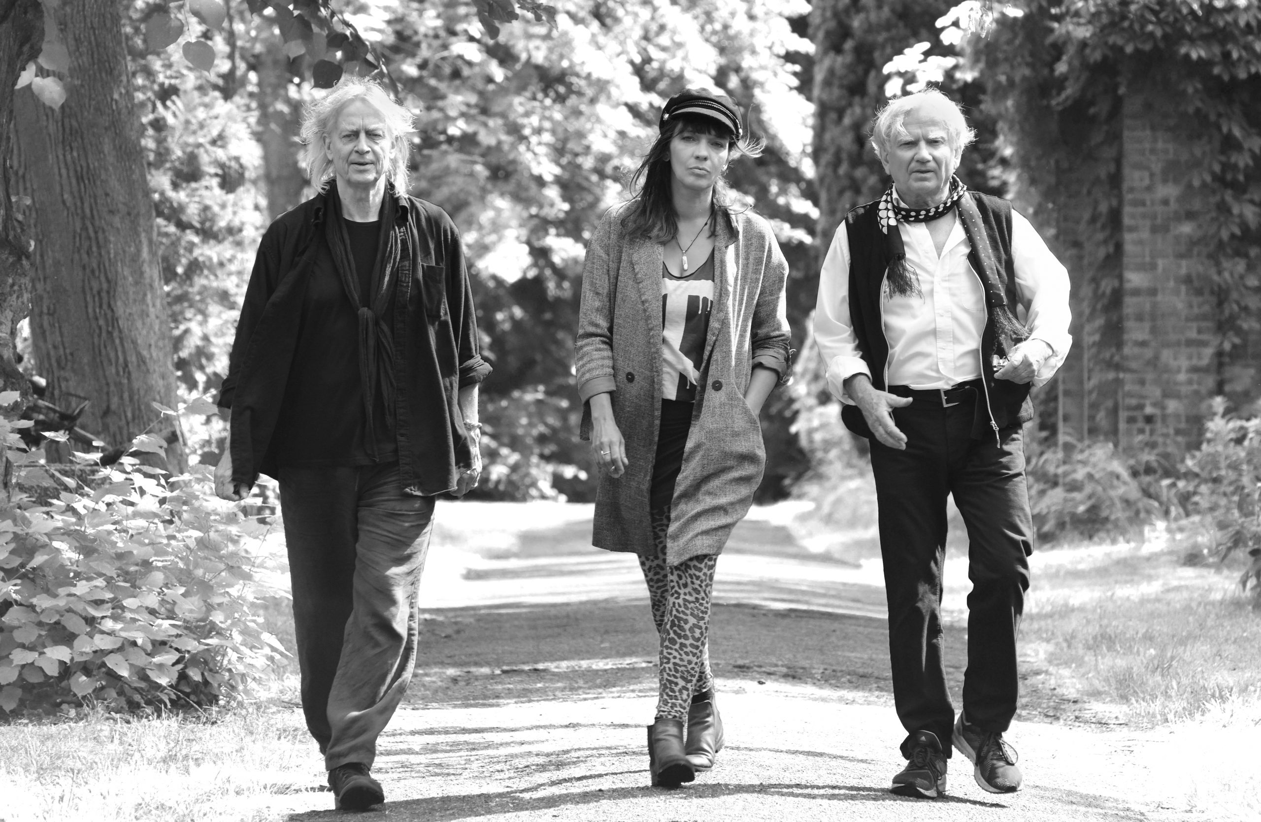 Drei Personen gehen auf einem von Bäumen gesäumten Weg, zwei Männer und eine Frau in legerer Kleidung.