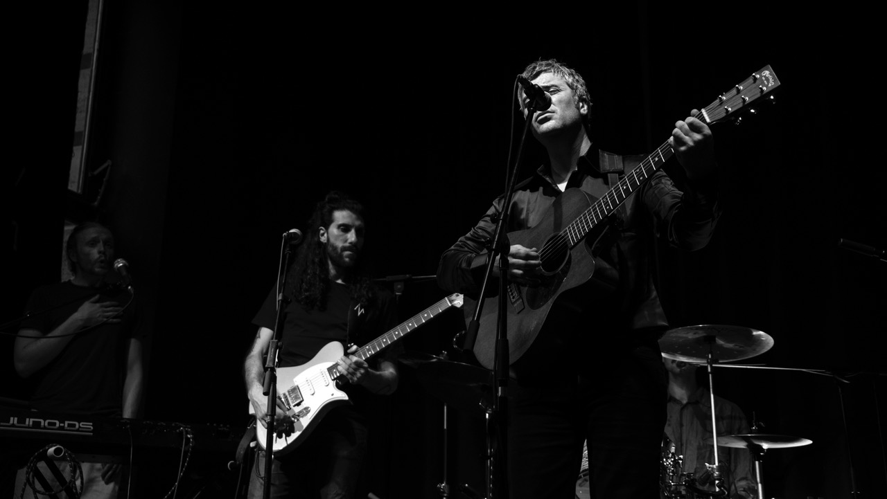 Drei Musiker auf einer Bühne, einer spielt akustische Gitarre, einer E-Gitarre, der dritte steht neben einem Keyboard