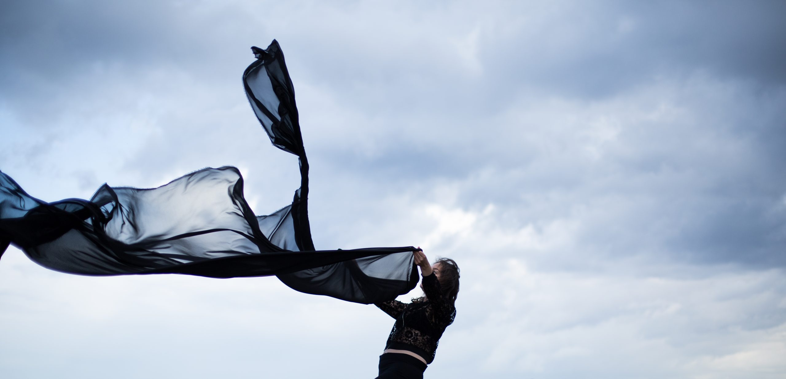 Eine schwarzgekleidete Person hält ein großes schwarzes Tuch in den Händen, das vom Wind verweht wird. Die Person steht vor einem blau grauen Himmel.